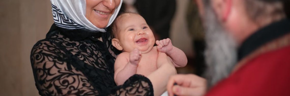 A smiling baby being baptized in a church, held by a woman with a headscarf.