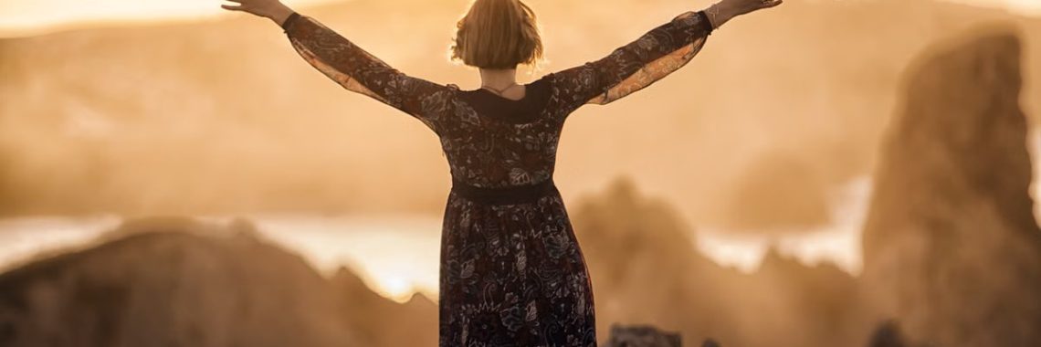 A woman stands with open arms on a mountain at sunset, embracing nature's beauty.